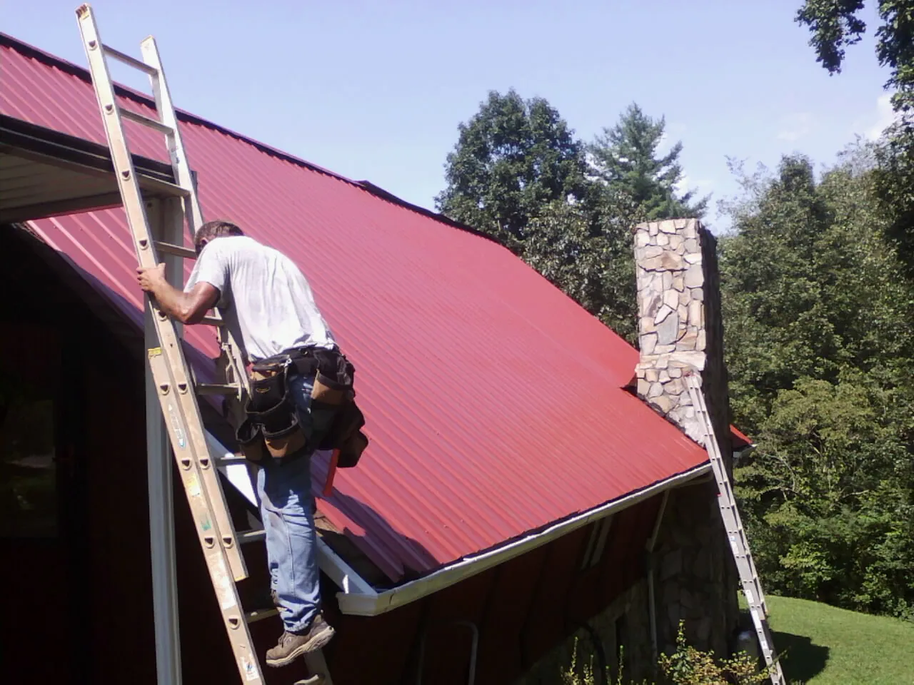 Roofer on a ladder beside a red metal roof and stone chimney — Soffit & Fascia Repair by Roofing Brothers in Dugway