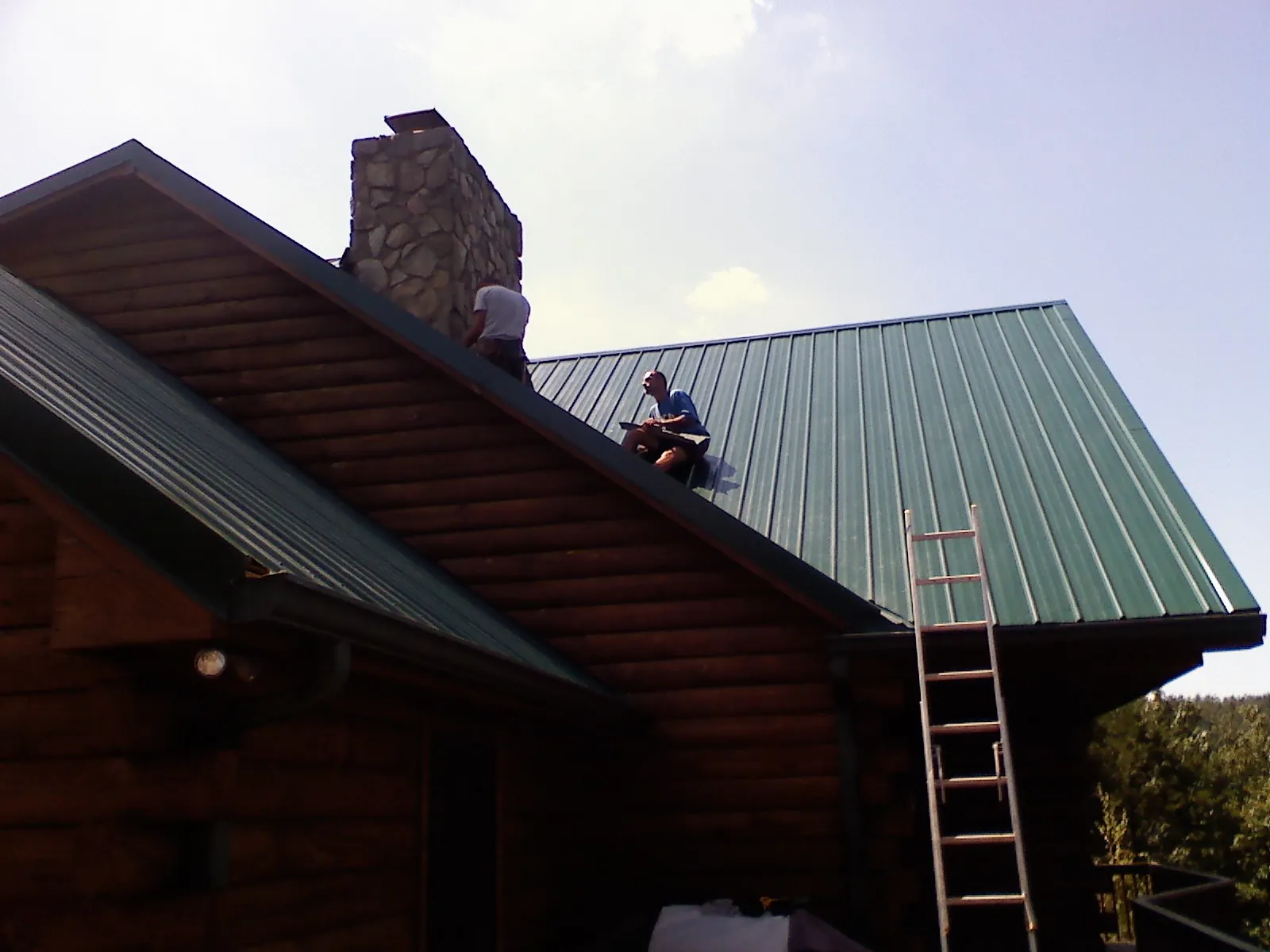 Roofers installing green metal panels beside a stone chimney on a log home in Dugway